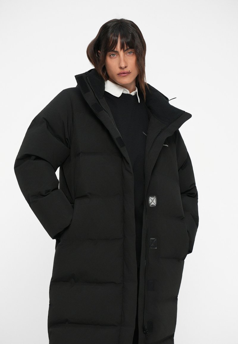 Black puffer coat featuring a high collar, large pockets, and a zip closure. The fabric appears smooth and insulated with quilted sections.