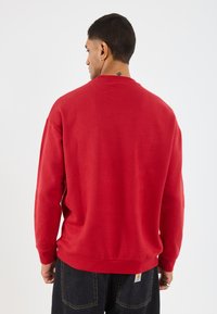 Rode katoenen sweatshirt met lange mouwen en een ronde halslijn. Heeft een ontspannen pasvorm en een boord aan de onderkant; zichtbaar Carhartt-label aan de achterkant.