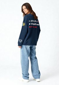 Felpa navy con testo grafico e ricamo a sole, vestibilità comoda, colletto e polsini a coste, abbinata a jeans baggy azzurri e sneakers.