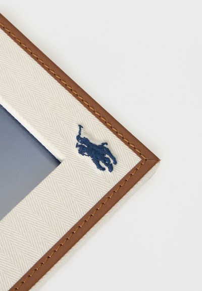 Γωνία ενός λευκού πορτοφολιού από υφή υφάσματος με καφέ δερμάτινη επένδυση και μικρό κεντημένο λογότυπο παίκτη πόλο σε ναυτικό μπλε χρώμα.