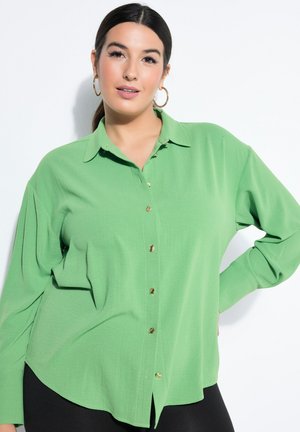 Kvinde med mørkt hår iført en grøn skjorte med knapper og guld øreringe i form af ringe, poserer med den ene hånd på hoften mod en hvid baggrund.