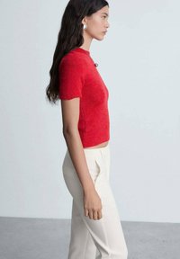 Top roșu cu mânecă scurtă, din tricot cu textură ribbed, combinat cu pantaloni albi, croiți; modelul stă de profil pe un fundal deschis.