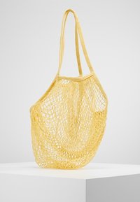 Gele nettas met lange hengsels, gemaakt van geweven materiaal; heeft een lichtgewicht, open ontwerp dat ideaal is voor het dragen van spullen.