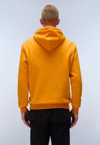Geel hoodie met een gestructureerde afwerking, kangoeroezak en ribgebreide boorden. Zichtbare capuchon aan de achterkant, gecombineerd met zwarte broek.
