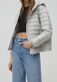Femme portant une doudoune gris clair à capuche sur un top court noir et un jean bleu taille haute, main dans la poche.