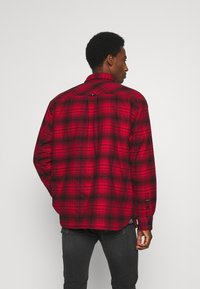 Red and black plaid flannel shirt with long sleeves, button-up closure, and a rounded hem, featuring a contrasting inner fabric at the collar.