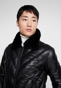 Zwarte leren jas met een gequilte patroon, een faux fur kraag en een ritssluiting aan de voorkant. Het ontwerp heeft schuine naden voor extra textuur.