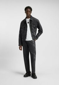 Schwarze Denimjacke mit vorderen Taschen, kombiniert mit einem weißen T-Shirt und schwarzen Hosen mit diagonalem Muster sowie schwarzen Schuhen.
