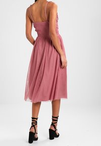 Vaaleanpunainen midi-mekko, jossa on smokkibodice ja laskeutuva hame. Mustat nauha-ankkuriset matalat korot. Sileä kangas kerroksellisella tekstuurilla.