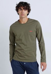 T-shirt à manches longues vert olive en coton, avec un col rond et un petit logo rouge sur la poitrine. Assorti à un pantalon bleu marine.