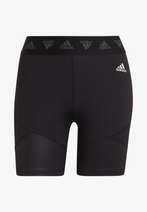 Atletisk person iført sorte Adidas kompressionsshorts og sorte løbesko, stående med benene spredt på hvid baggrund.