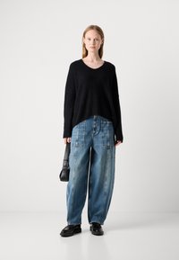 Suéter negro texturizado con escote en V, combinado con jeans de mezclilla de gran tamaño en un tono azul claro con costuras llamativas. La modelo sostiene un pequeño bolso negro.