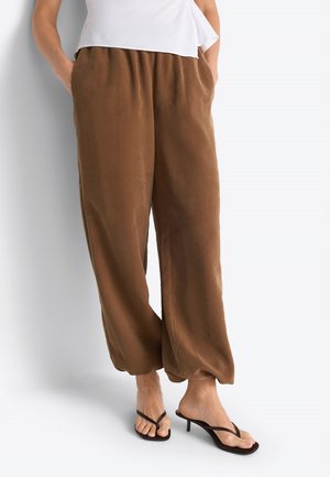 Donna che indossa pantaloni marroni larghi con polsini elastici e sandali neri con tacco, in piedi contro uno sfondo chiaro e uniforme.