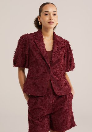 Vrouw die een textuurrijke bordeauxrode korte mouw blazer en bijpassende shorts draagt, met gouden oorringen en handen in de zakken, neutrale achtergrond.