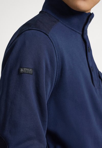 Temno modra polo majica z visokim ovratnikom, ima tri gumbe, teksturirano tkanino in kontrastno ramensko obliž z logotipom.