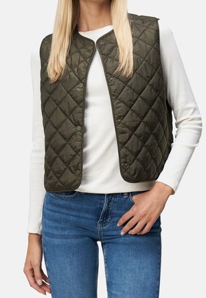 Kobieta w oliwkowozielonej pikowanej kamizelce bez rękawów na białej koszuli z długim rękawem i w niebieskich dżinsach, stojąca z jedną ręką na biodrze.