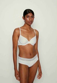 Ženska z temnimi lasmi, oblečena v belo modrček z tankimi naramnicami in ustrezno belo spodnje perilo z čipkastim pasom, ki stoji ob enobarvni svetli ozadju.