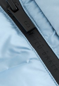 Ljusblått tyg med en slät textur, med en svart dragkedja och den präglade namnet "TED BAKER" på dragkedjerepet.