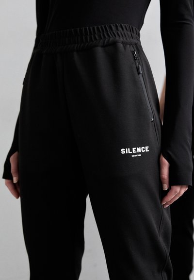 Svarta sweatpants med elastiskt midjeband, en ficka med dragkedja och en vit logotyp som säger "SILENCE" på benet. Mjukt material.