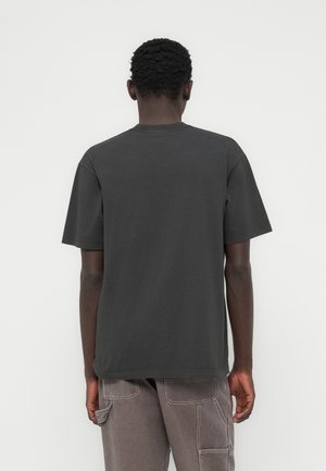 Person med kort krøllet hår iført en ensfarvet mørkegrå t-shirt og lysegrå bukser, set bagfra mod en hvid baggrund.