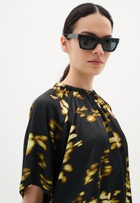 Blusa nera con stampa floreale astratta in giallo e oro, scollo arricciato, maniche corte e texture liscia e fluida. Occhiali da sole neri.