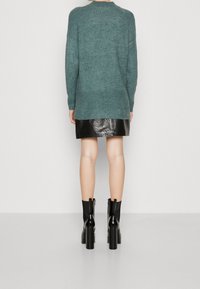 Pull en tricot teal avec un ourlet et des manches côtelés, associé à une jupe en cuir noir et des bottines à talons noires, vu de dos.