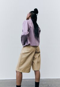Helllila strukturierte Bluse mit weiten Ärmeln, kombiniert mit beigen knielangen Shorts, die über seitliche Nähte und Gesäßtaschen verfügen. Sichtbare schwarze Socken.