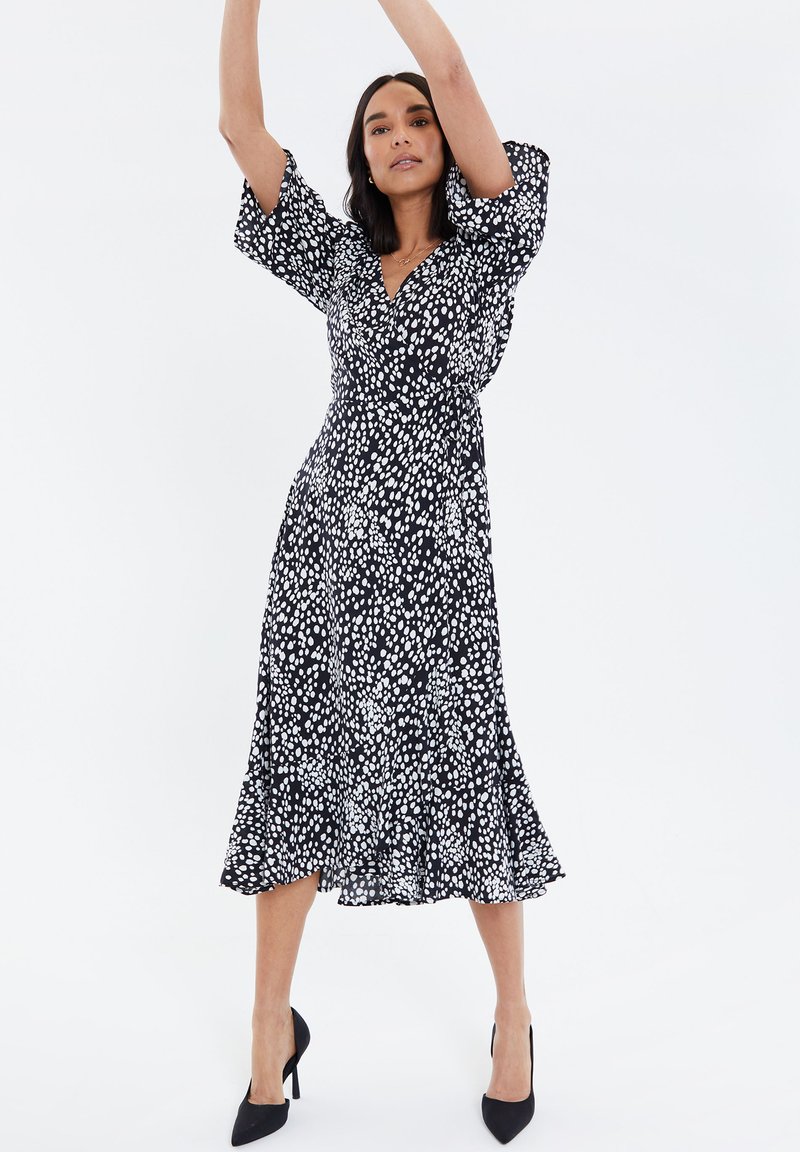 Vestido negro con lunares blancos, mangas cortas abullonadas, escote en v, cintura ajustada, dobladillo acampanado y un borde con volantes en la parte inferior.