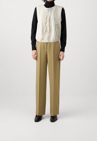 Béžová vesta bez rukávů s kulatým výstřihem, předním zipem a bočními kapsami, doplněná vysokým pasem a rovnými hnědými kalhotami a černou obuví.