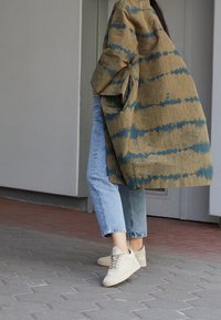 Długa, wzorzysta kurtka w oliwkowym i niebieskim kolorze, zestawiona z jasnoniebieskimi dżinsami i beżowymi sneakersami. Teksturowana tkanina z szerokimi rękawami i bocznymi kieszeniami.