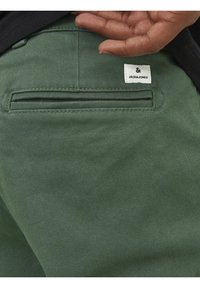 Zielone bawełniane spodnie mają tylną kieszeń z zamkiem błyskawicznym oraz białą metkę z nazwą marki "JACK & JONES" wszytą na niej.