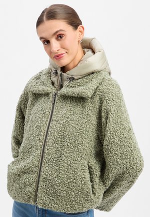 Woman with tied-back hair wearing a green textured zip-up jacket over a beige hooded layer, hands in pockets, smiling gently against a plain background.