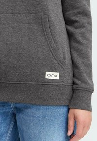 Main droite à côté de la poche d'un sweat-shirt gris foncé avec une étiquette "OXMO", portant un jean bleu.