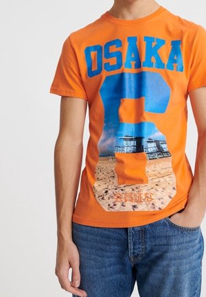 T-shirt orange vif avec le texte "OSAKA" et un grand chiffre 8 représentant une scène de plage avec des postes de sauveteurs et du sable.