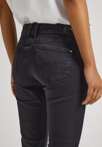 A fekete farmernadrág slim fit szabású, hátsó zsebei diszkrét varrással készültek, sima textúrával rendelkeznek, és finoman fakó hatásúak.