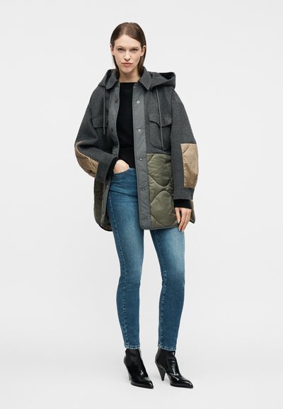 Γυναίκα που φοράει μια σακάκι παζλ με κουκούλα και ραμμένα μπαλώματα σε ελιά και μπεζ, μαύρο πουλόβερ, στενά μπλε τζιν και μαύρες μπότες αστραγάλου με τακούνι.