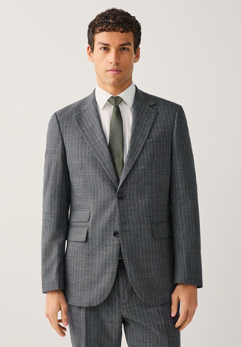 Homme portant un costume gris à fines rayures avec deux boutons, une chemise blanche et une cravate verte à pois, debout devant un fond clair uni.