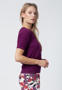 Suéter de punto corto de manga corta en color púrpura con un patrón de diamante texturizado. Combinado con pantalones de estampado floral en colores blanco, rojo y púrpura.