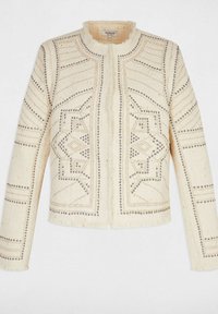 Giacca color crema con un tessuto texturizzato e intrecciato, caratterizzata da motivi geometrici delineati da borchie metalliche e un orlo frangiato.