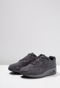 Šedé semišové tenisky s polstrovaným límcem, kulatou špičkou a šněrováním. Mají černou gumovou podrážku a logo Nike na boku.