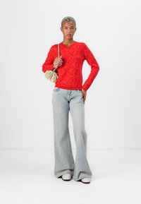 Maglione lavorato a maglia con motivo floreale rosso e scollatura a V, abbinato a jeans azzurri a zampa d'elefante e sneakers bianche, mentre tiene una piccola borsa crema.