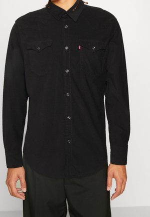 Mann trägt ein schwarzes, langärmeliges Hemd mit zwei Brusttaschen und Metall-Druckknöpfen und steht vor einem schlichten Hintergrund.