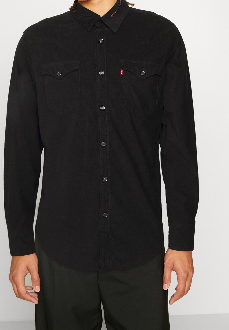 Homme portant une chemise noire à manches longues boutonnée avec deux poches poitrine et des boutons-pression métalliques, debout devant un fond uni.