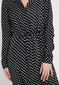 Svart klänning med prickar i lätt tyg. Har en v-ringning, långa ärmar, midjeband och en flödande design med vita cirkulära mönster.