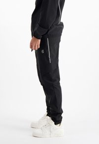 Zwarte sportieve broek met een gladde textuur, zijzakken met rits en reflecterende accenten. Gecombineerd met witte sneakers.