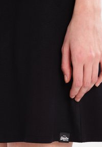 Musta puuvillamekko, jolla on sileä pinta, jossa on helman viimeistely ja pieni brändilogo alareunassa. Käsi lepottelee kevyesti kankaan päällä.