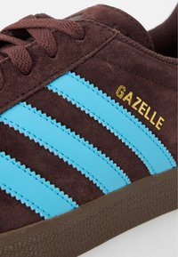 Brązowy zamszowy sneaker z trzema niebieskimi syntetycznymi paskami. Złote logo "GAZELLE", brązowa teksturowana gumowa podeszwa oraz okrągłe sznurowadła.