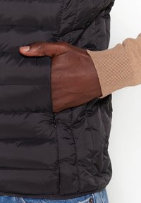 Doudoune sans manches noire à motif matelassé horizontal, avec une poche zippée. Détail de poignet côtelé marron clair visible au niveau du poignet.