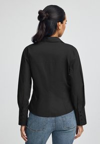 Kvinna med mörkt hår i en knut, iklädd en tight svart skjorta med knappar och blå jeans, sedd bakifrån mot en enfärgad bakgrund.