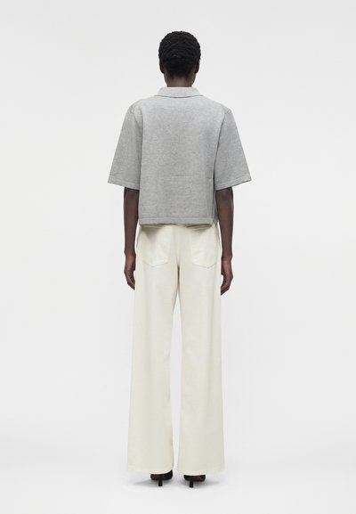 Άτομο που στέκεται με την πλάτη στην κάμερα, φοράει γκρι μπλουζάκι με κοντά μανίκια, φαρδιά κρεμ παντελόνια και μαύρα παπούτσια με τακούνι σε λευκό φόντο.